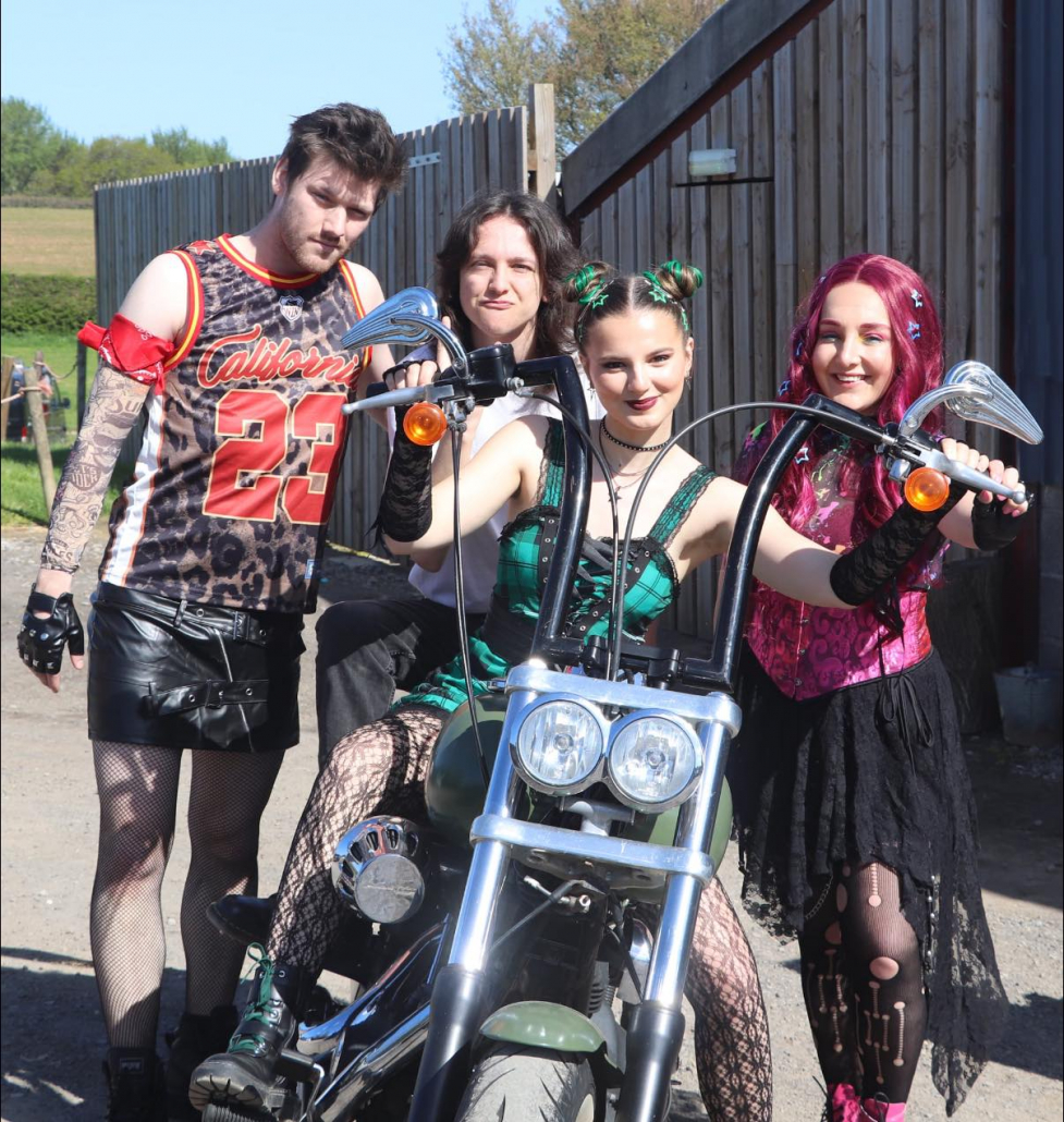 the cast of "We Will Rock You" in costume for a promotional photo-shoot. "Scaramouche" is astride a motorbike with other characters around her.